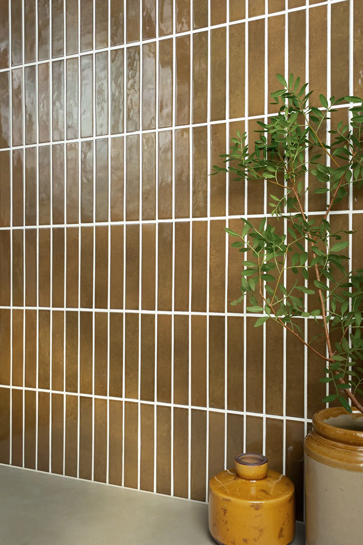 Slim, glossy toffee brown metro wall tiles laid vertically with white grout, styled with green foliage and ochre ceramic accessories