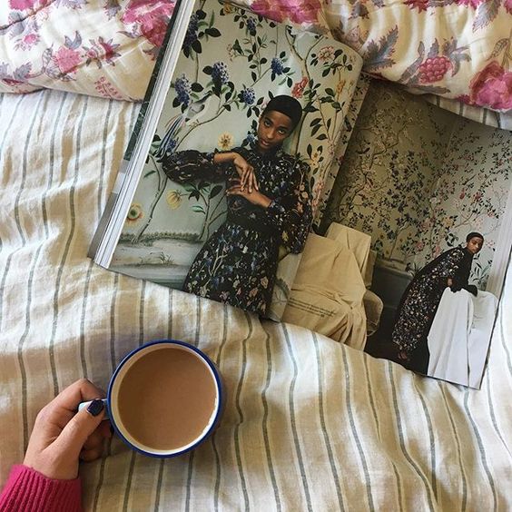 We love interiors magazines! Lifestyle flatlay of a reader in bed holding a cup of tea and reading an open magazine.