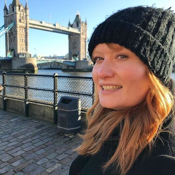 The top UK interior stylists you should be following. Picture of a red-head woman wearing a black hat in front of Tower Bridge in London