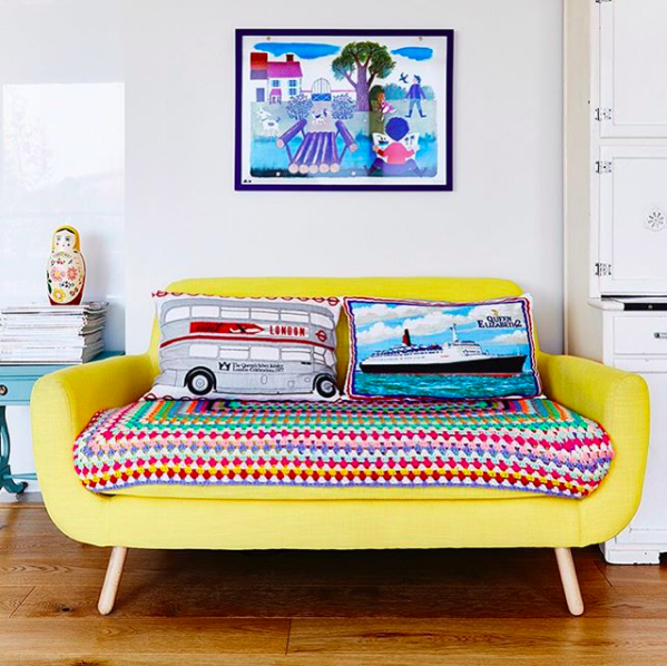 Brightly coloured yellow sofa styled in front of a colourful piece of wall art and with a brightly coloured throw and pillows.