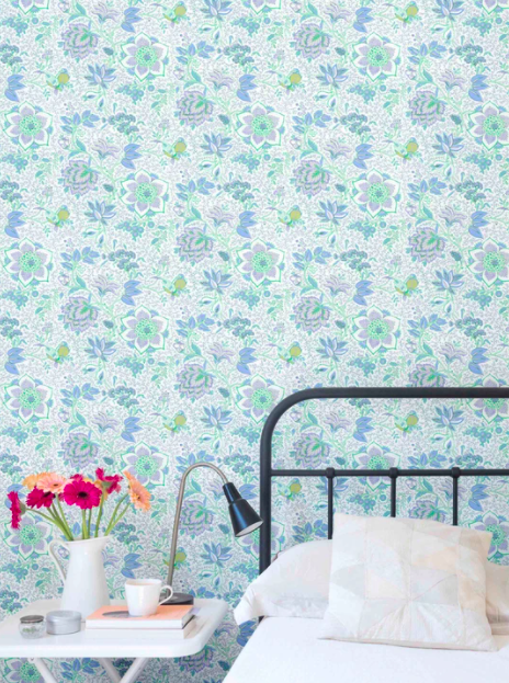 Green and blue floral wallpaper on a wall behind a black framed bed. A side table holds pink and yellow flowers in a white jug.