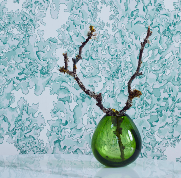 Wallpaper featuring green lichen styled behind a green glass vase holding a wooden branch.
